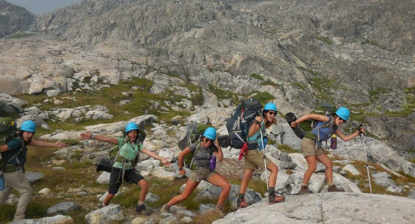 A group of people wearing backpacks and helmets make their way across a rocky landscape and make silly poses for the picture. 